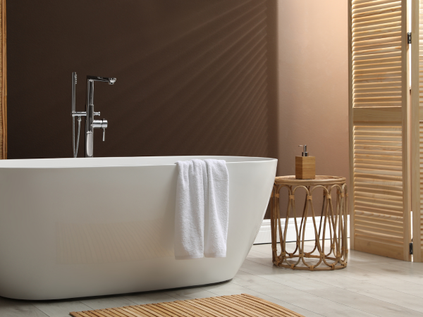 White freestanding bathtub next to rattan table against brown walls and wooden shutters