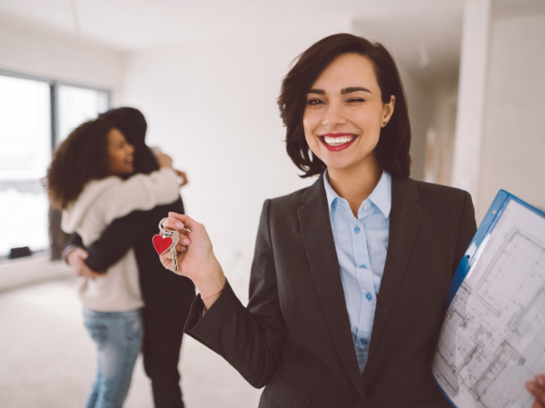 a young couple hugging after buying a house, while real estate agent holds their keys and smiles